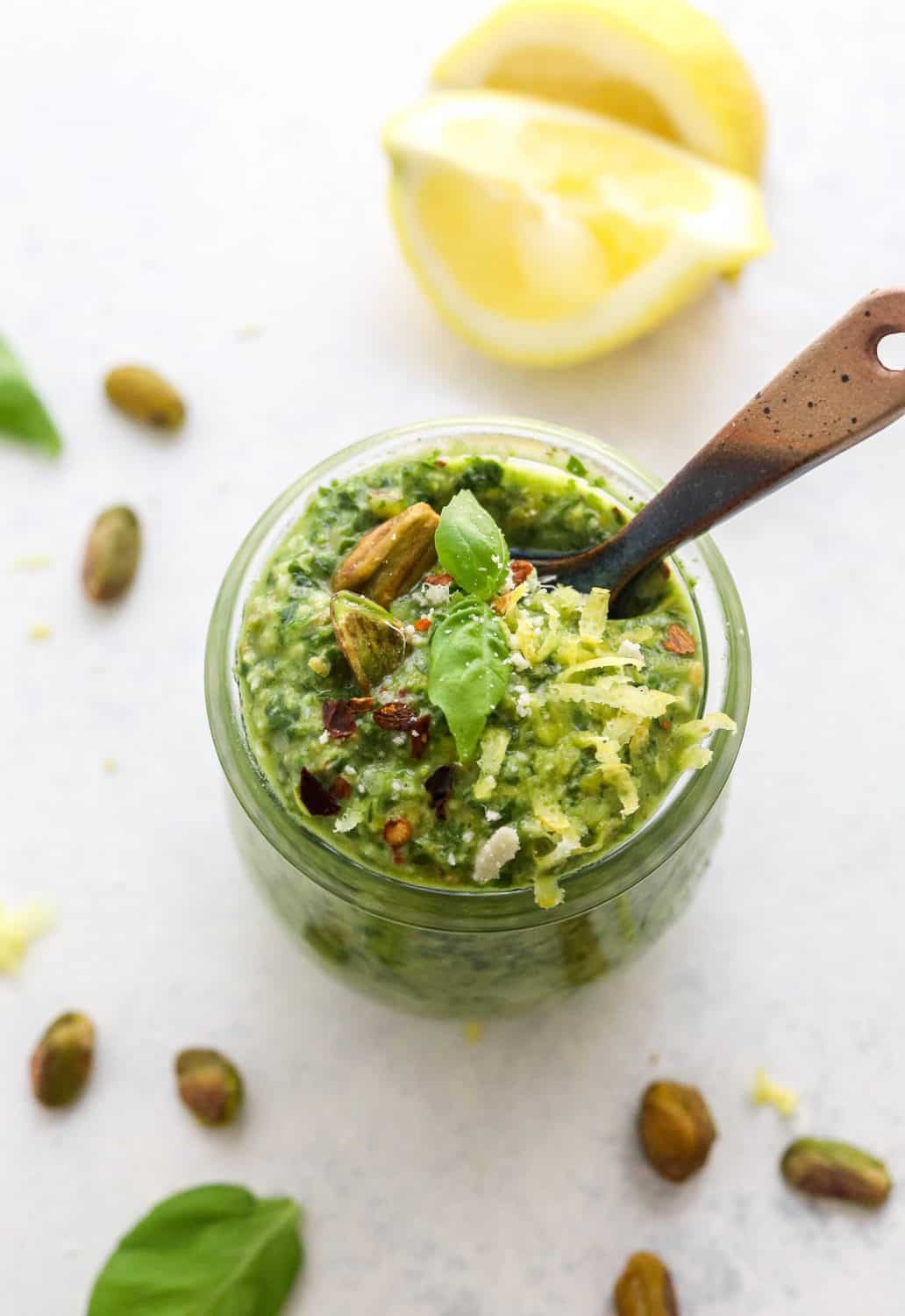Glass jar of green basil pesto with a blue spoon in it with some nuts and lemon around the  bottom of the jar