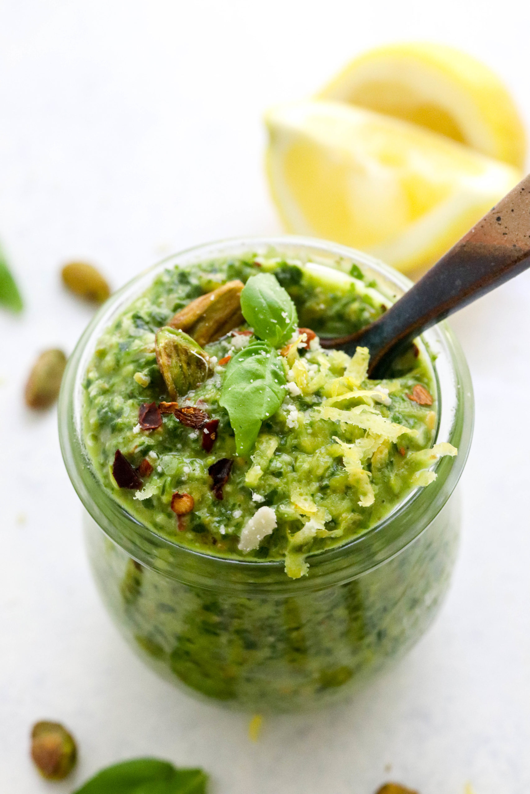 Round jar filled with green pesto topped with lemon zest and nuts with more sliced lemon and nuts behind it