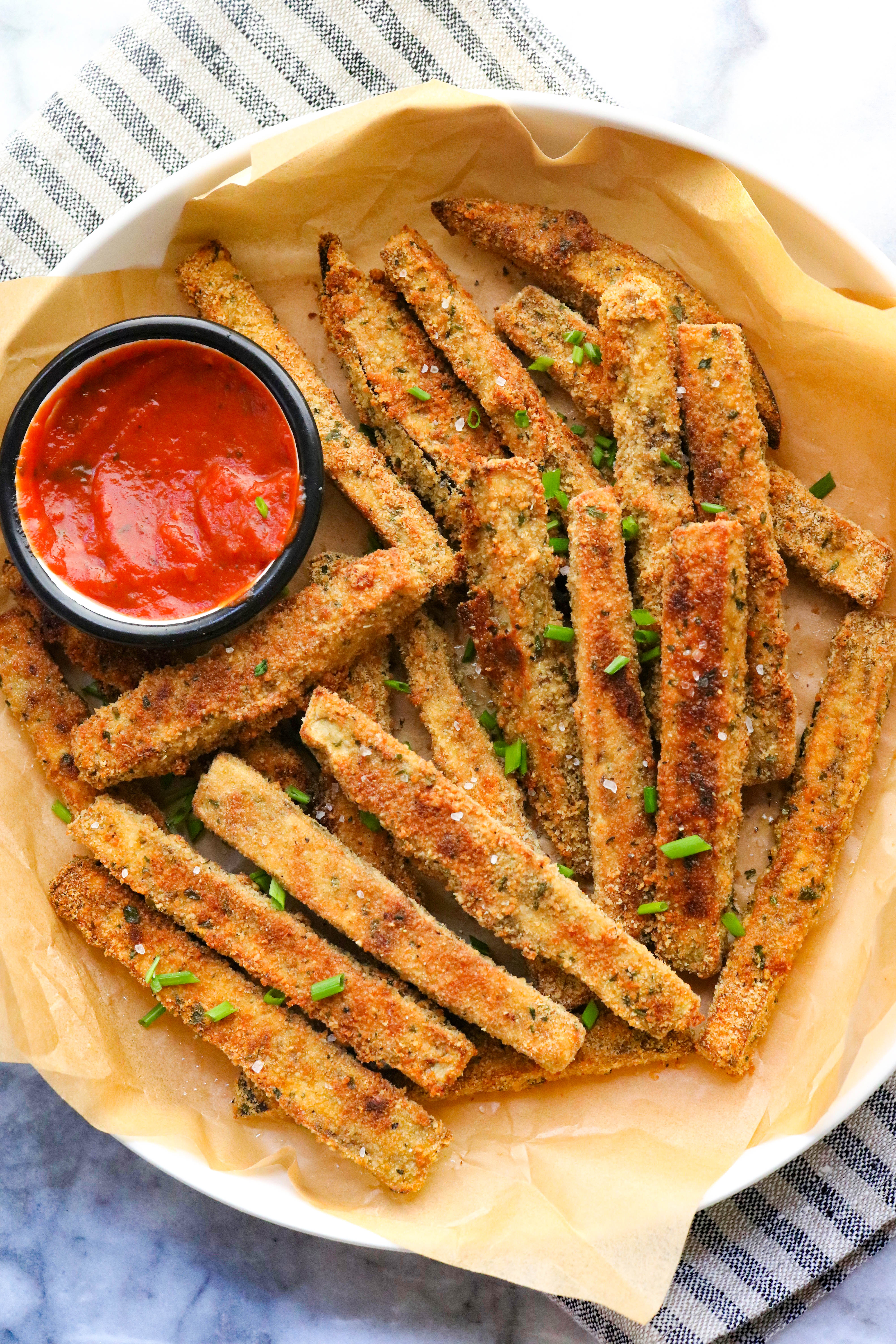 Several crispy breaded and baked fries on a plate with parchment paper and tomato sauce in a small bowl next to it. 