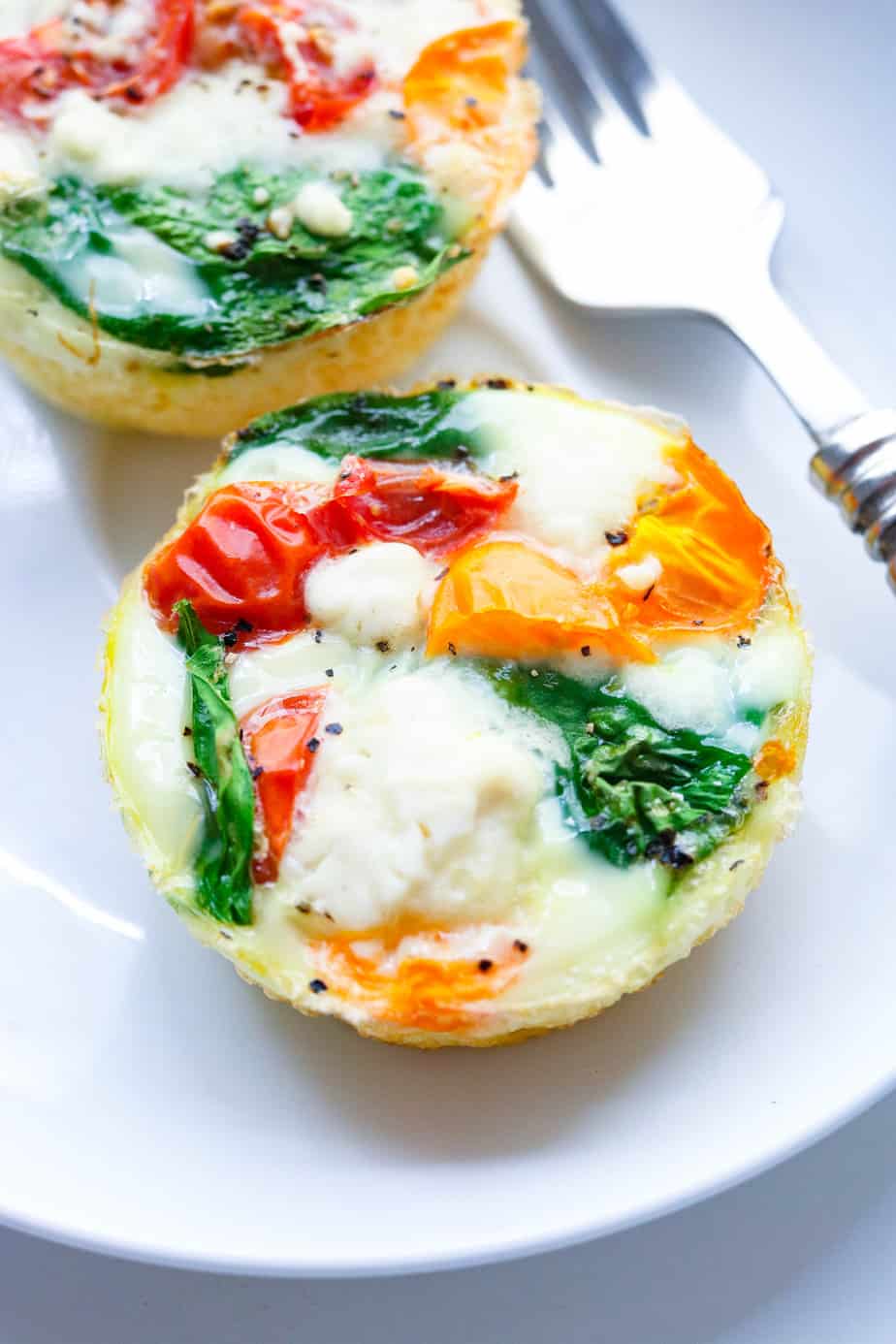Single egg white bite with red and green veggies in it on a white plate with a fork next to it. 