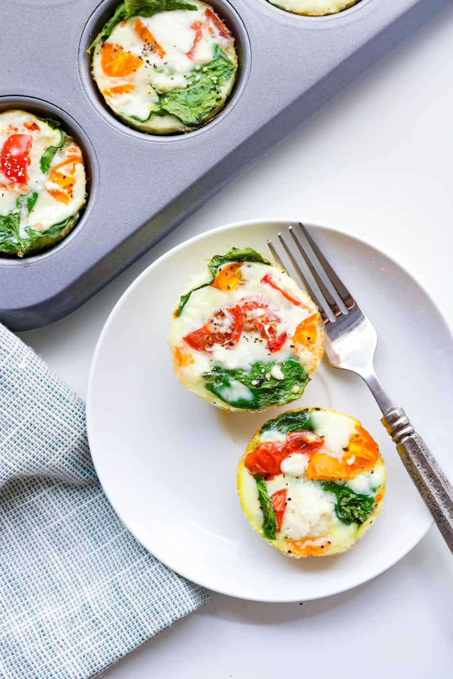 two egg muffins with veggies in them on a round white plate with a silver fork on the plate and a muffin pan with more egg muffins in it behind it
