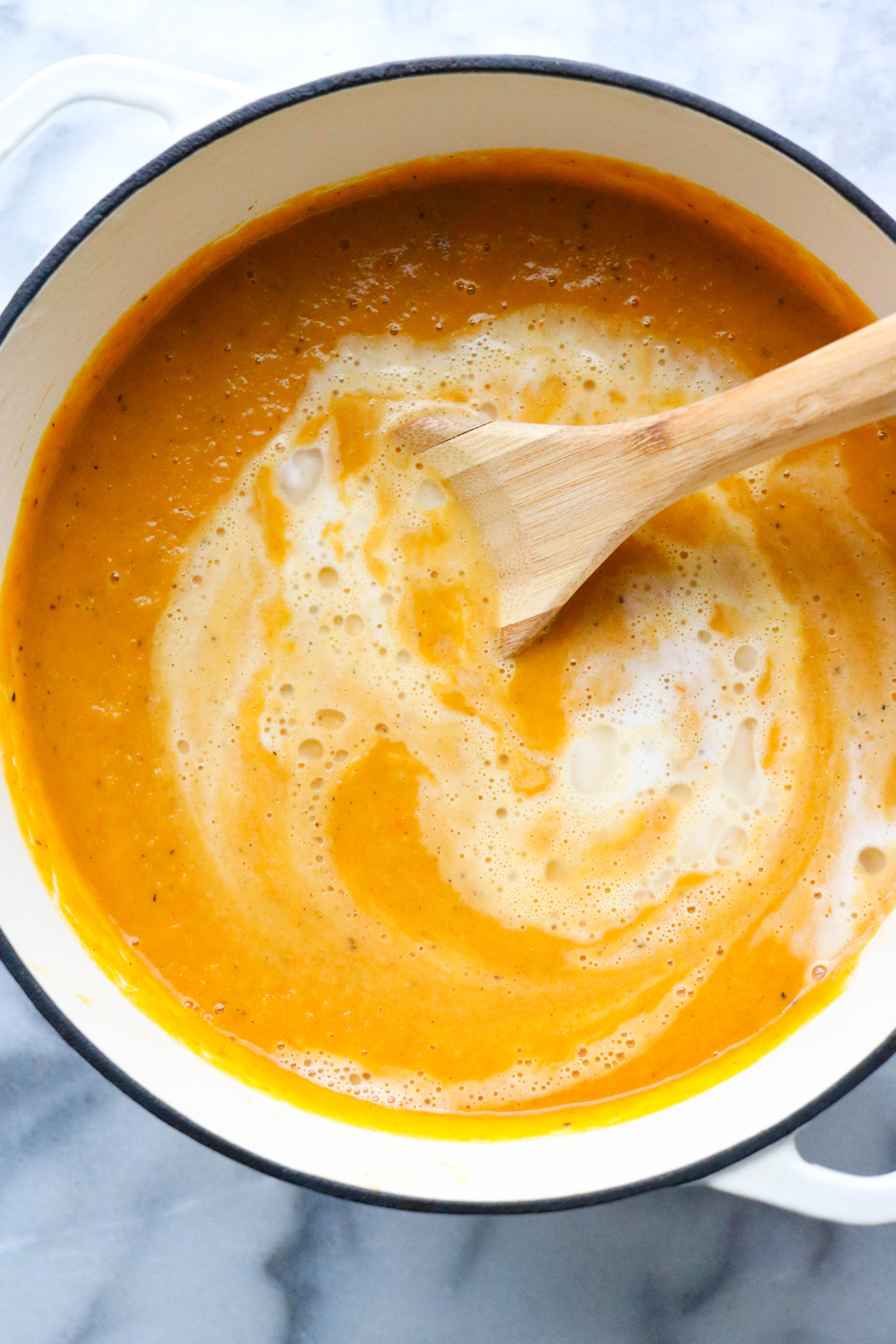Pot filled with smooth butternut squash soup with a swirl of cream in the soup and a wooden spoon in the bot on a marble surface. 