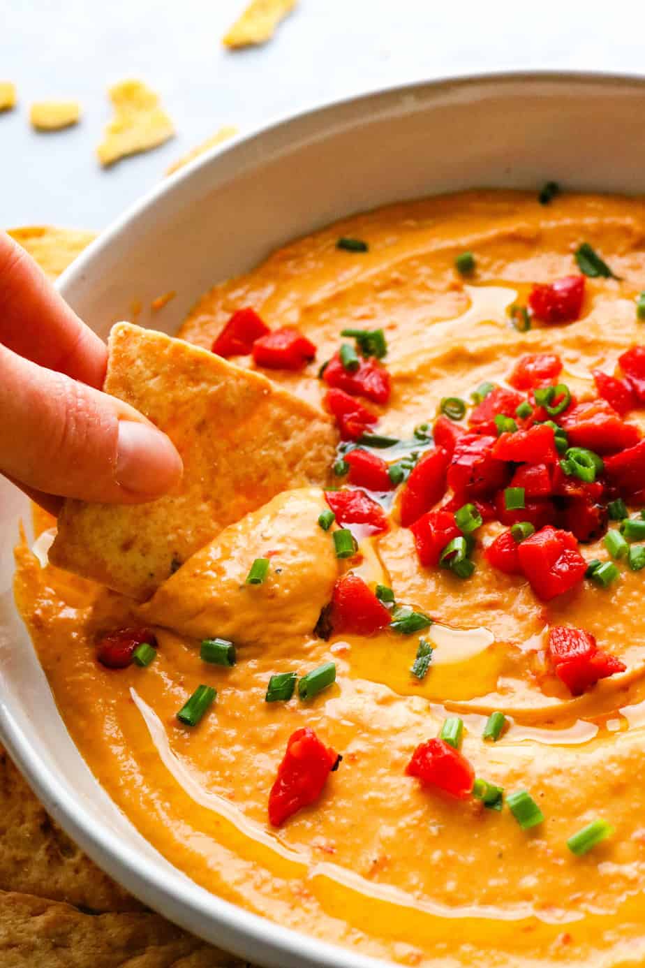 Roasted red pepper hummus in a bowl toped with chopped bell peppers and chives with a hand dipping a pita chip in the hummus. 