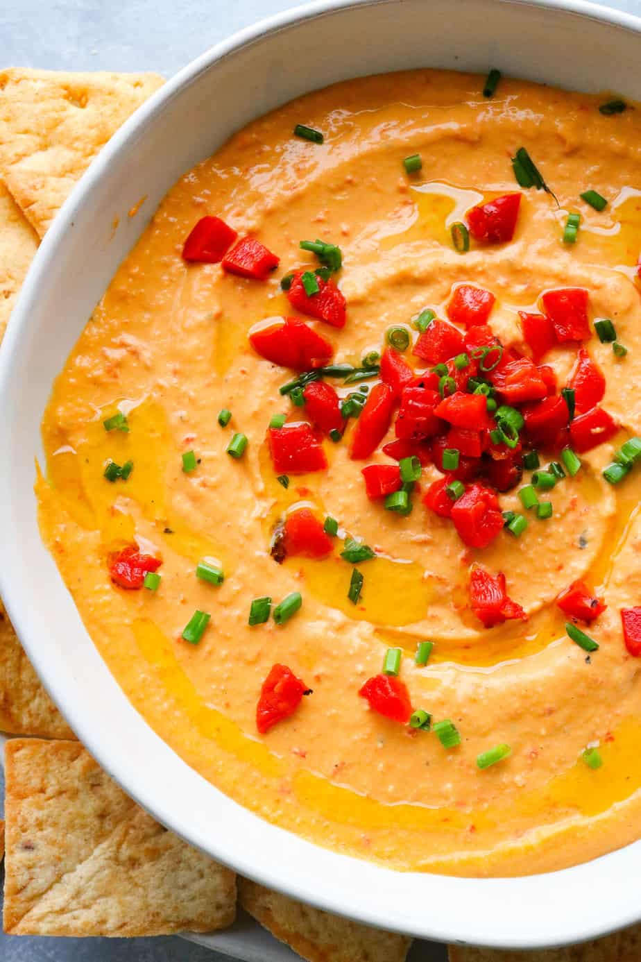 Bowl of orange hummus with chopped roasted red peppers and herbs on top and pita chips next to it. 