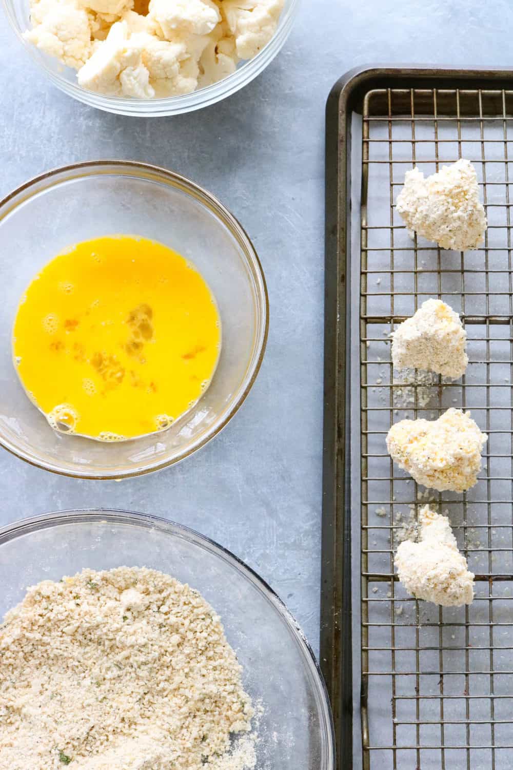 flour eggs and breadcrumbs in glass bowls next to a wire racked baking sheet