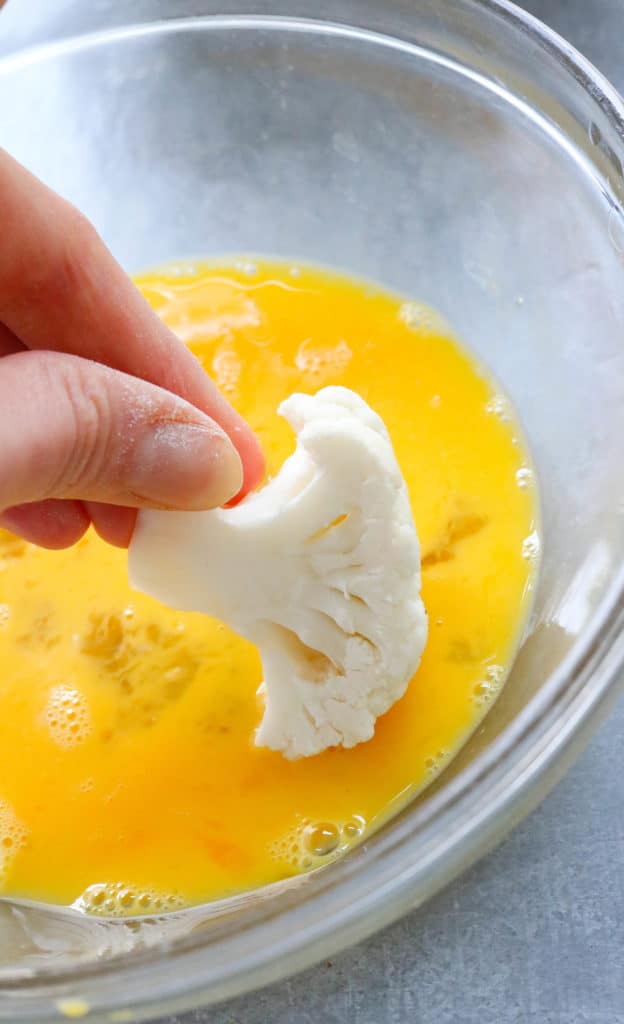 Uncooked piece of cauliflower being dipped into a bowl of whisked eggs. 
