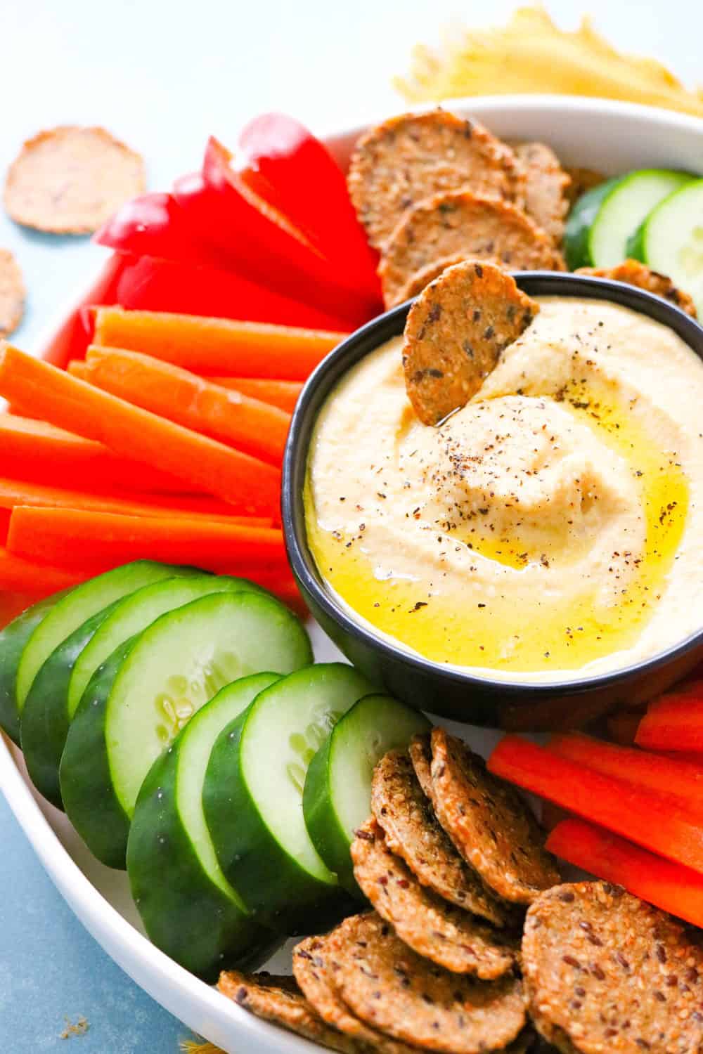 dipping cracker in homemade hummus on a platter with veggies and more crackers