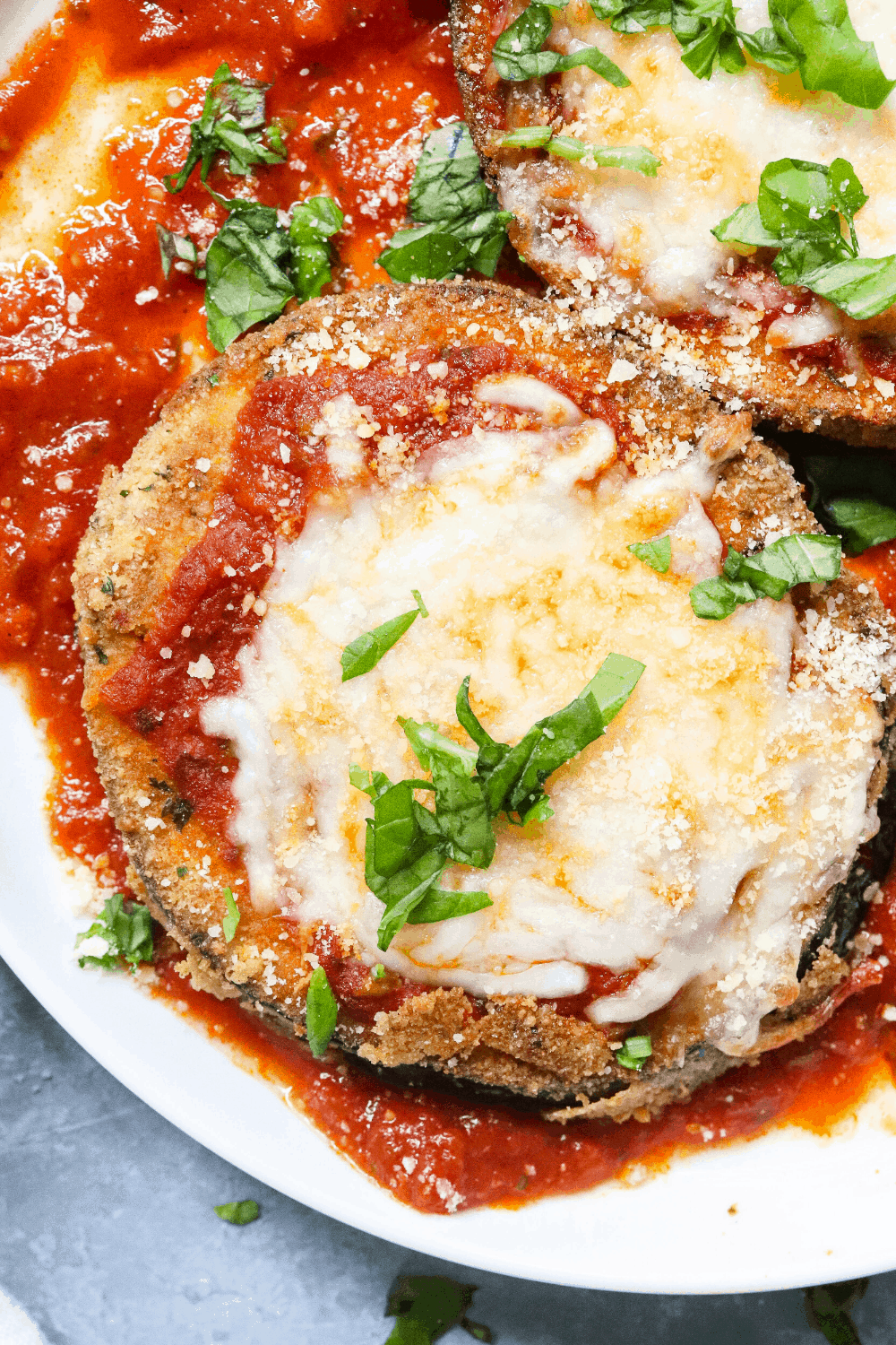 One piece of breaded eggplant parmesan on a plate with tomato sauce and sliced basil on top of it. 