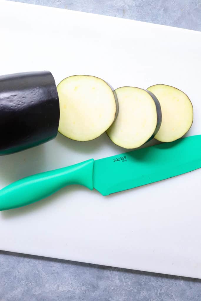 Sliced eggplant on a white cutting board with a green knife next to it