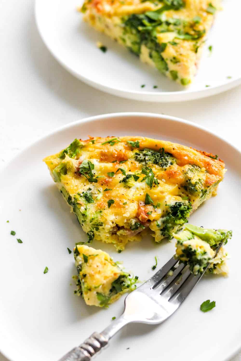 Ham and Broccoli quiche slice on a white plate with a fork cutting a piece out of it