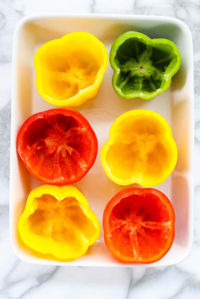 Red, yellow and green hollowed out bell peppers in a white baking dish. 