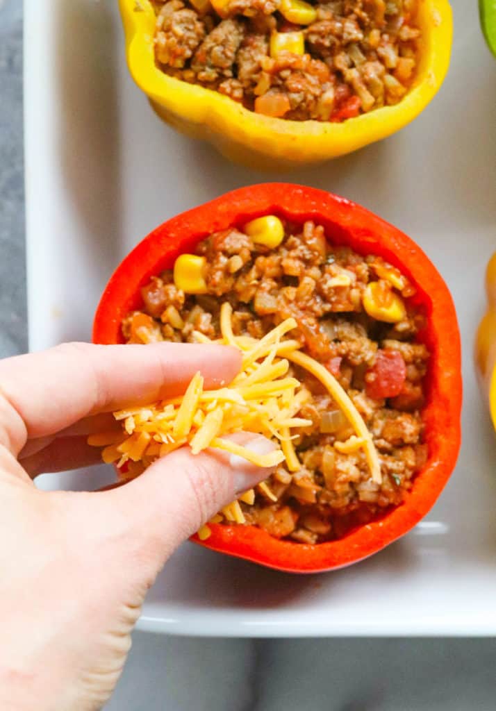 Hand sprinkling yellow cheddar cheese onto a red taco stuffed pepper with more stuffed peppers around it. 