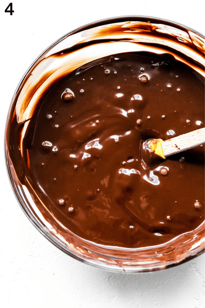 Glass bowl filled with melted dark chocolate with a rubber spatula in the bowl. 