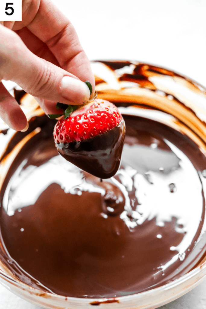 Hand dipping a strawberry into a bowl of melted dark chocolate. 