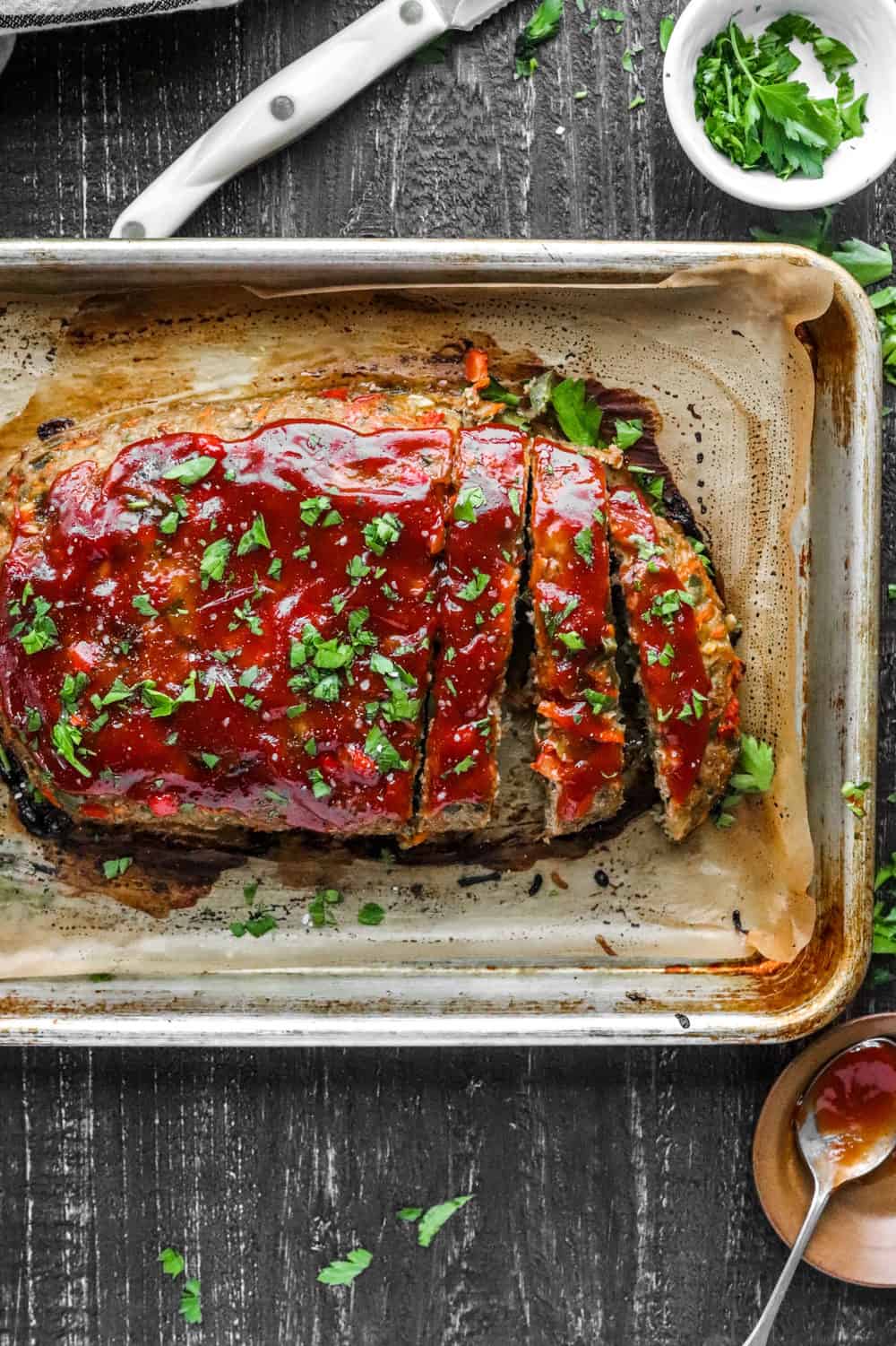 Cooked meatloaf cut into slices on a pan topped with red tomato sauce and parsley.