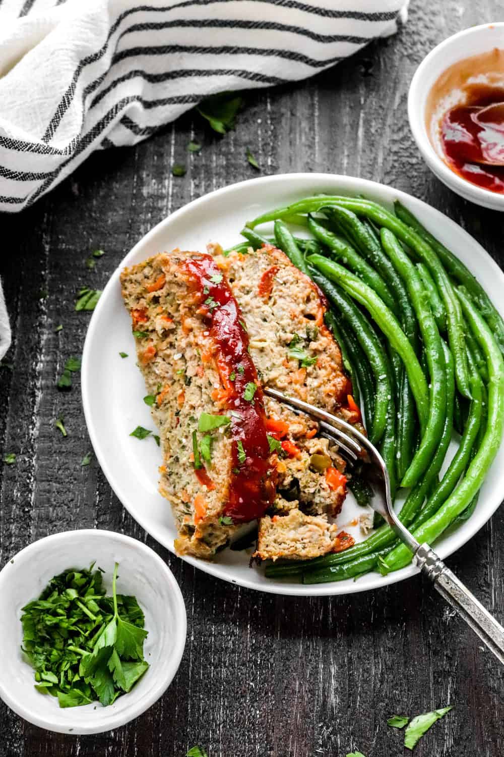 Green bean and sliced meatloaf on a round white plat with a fork in it with a stripped white and blue linen behind it an a bowl of chopped herbs in front of it. 