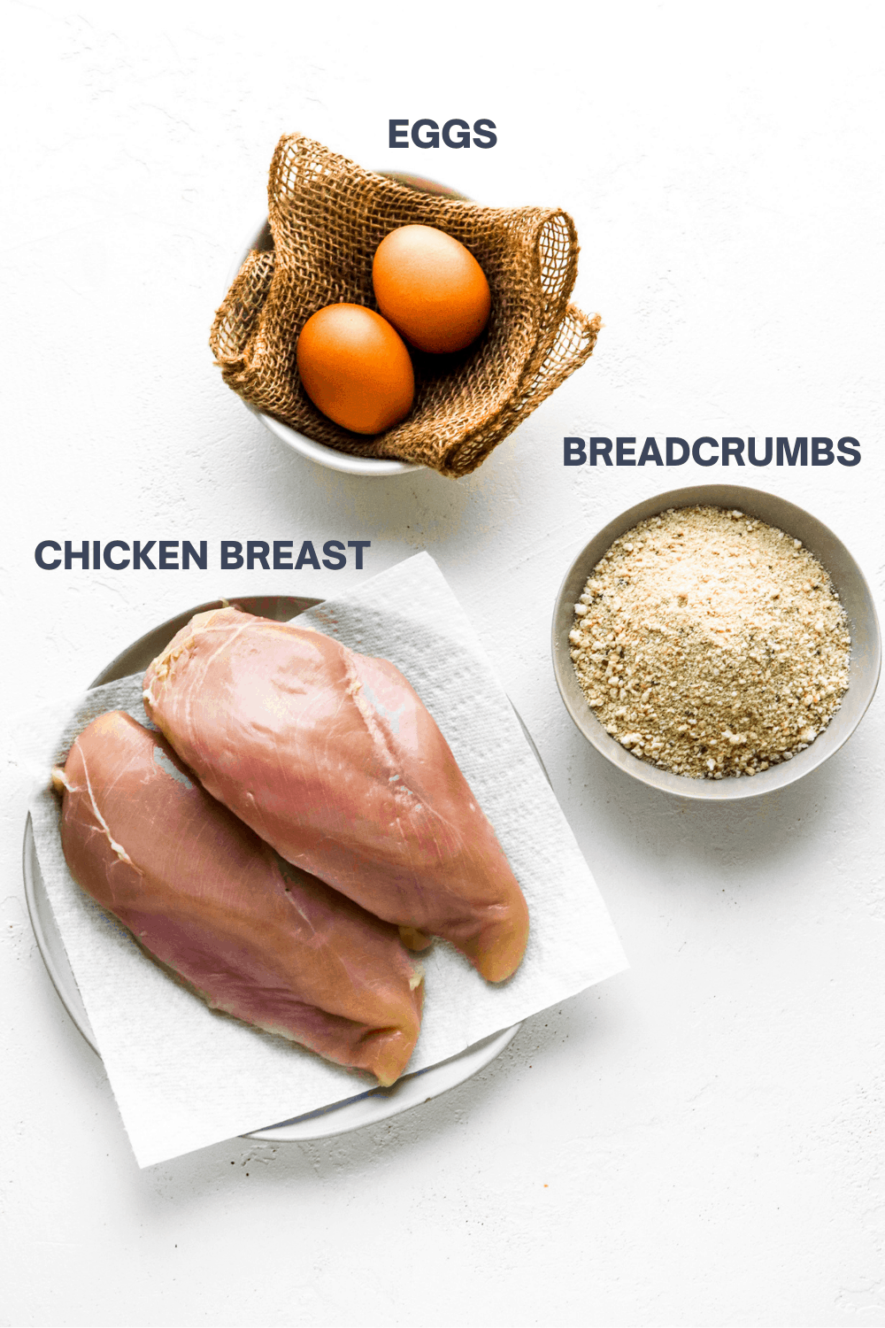 raw chicken breast, breadcrumbs and eggs on a white surface