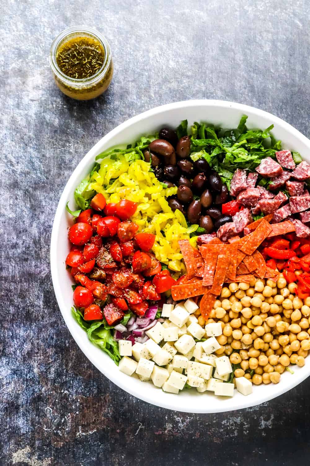 White salad bowl with lettuce in it topped with chopped tomatoes, olives, pepperoni, chickpeas and cheese