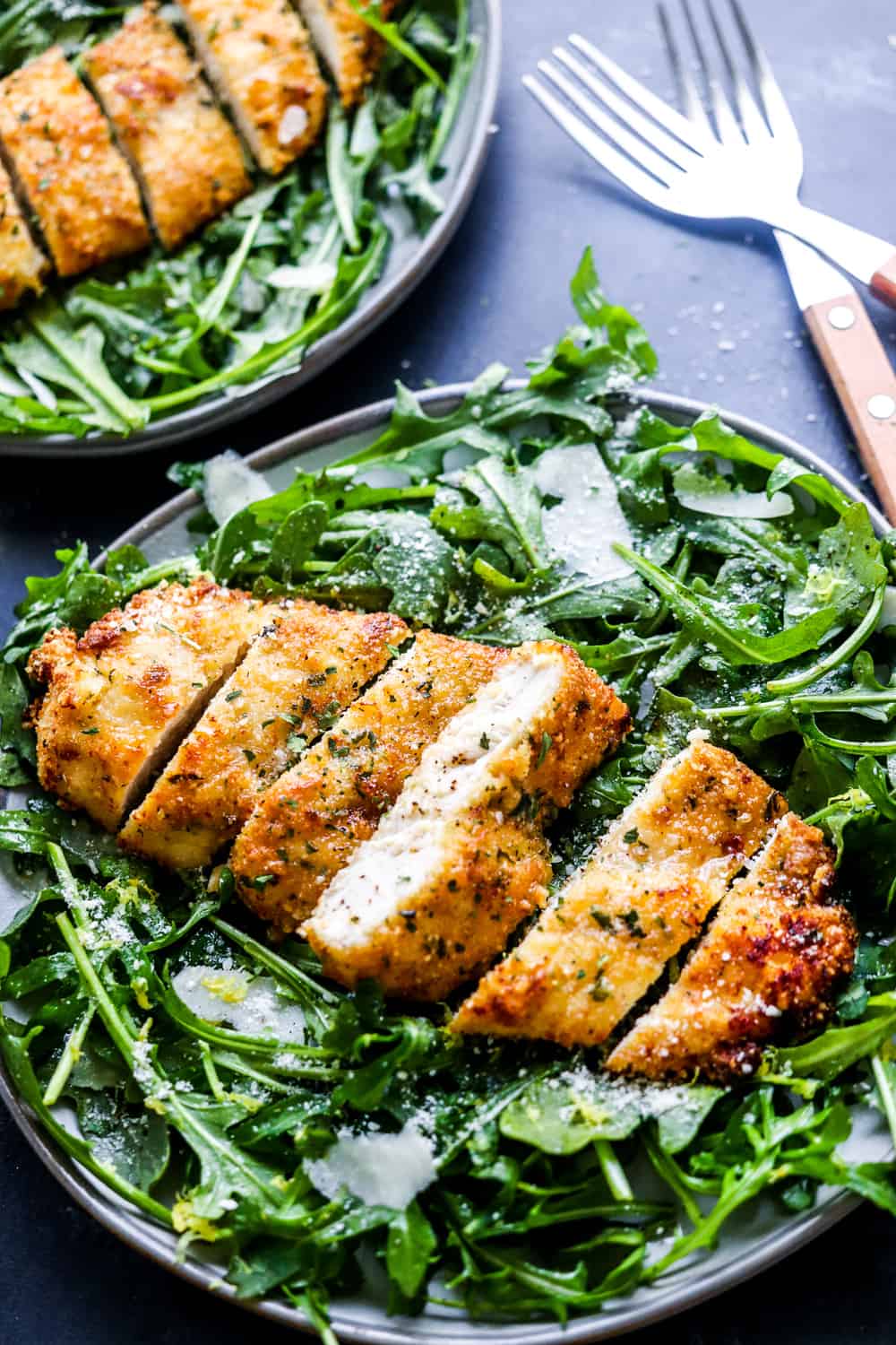 Plate filled with arugula salad topped with breaded chicken sliced with cutlery behind it. 