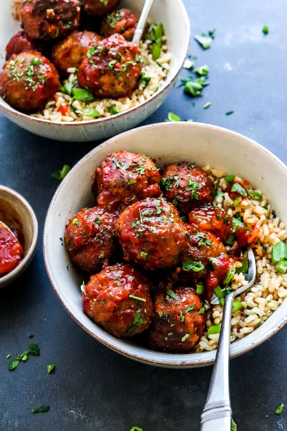 two grey round bowls filled with rice and meatballs covered in sauce with herbs on top of them and a fork sitting in one of the bowl. 