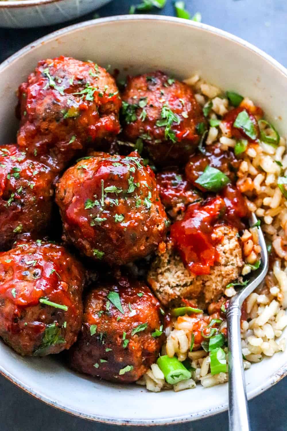 healthy turkey meatballs rolled in chili sauce in a bowl on top of brown rice with chopped scallions on top of it.