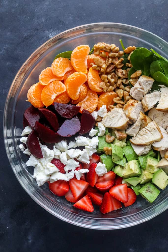 Round glass mixing bowl filled with roasted beets, orange slices, nuts, sliced strawberries, diced avocado, chopped chicken, and goat cheese on a dark blue counter