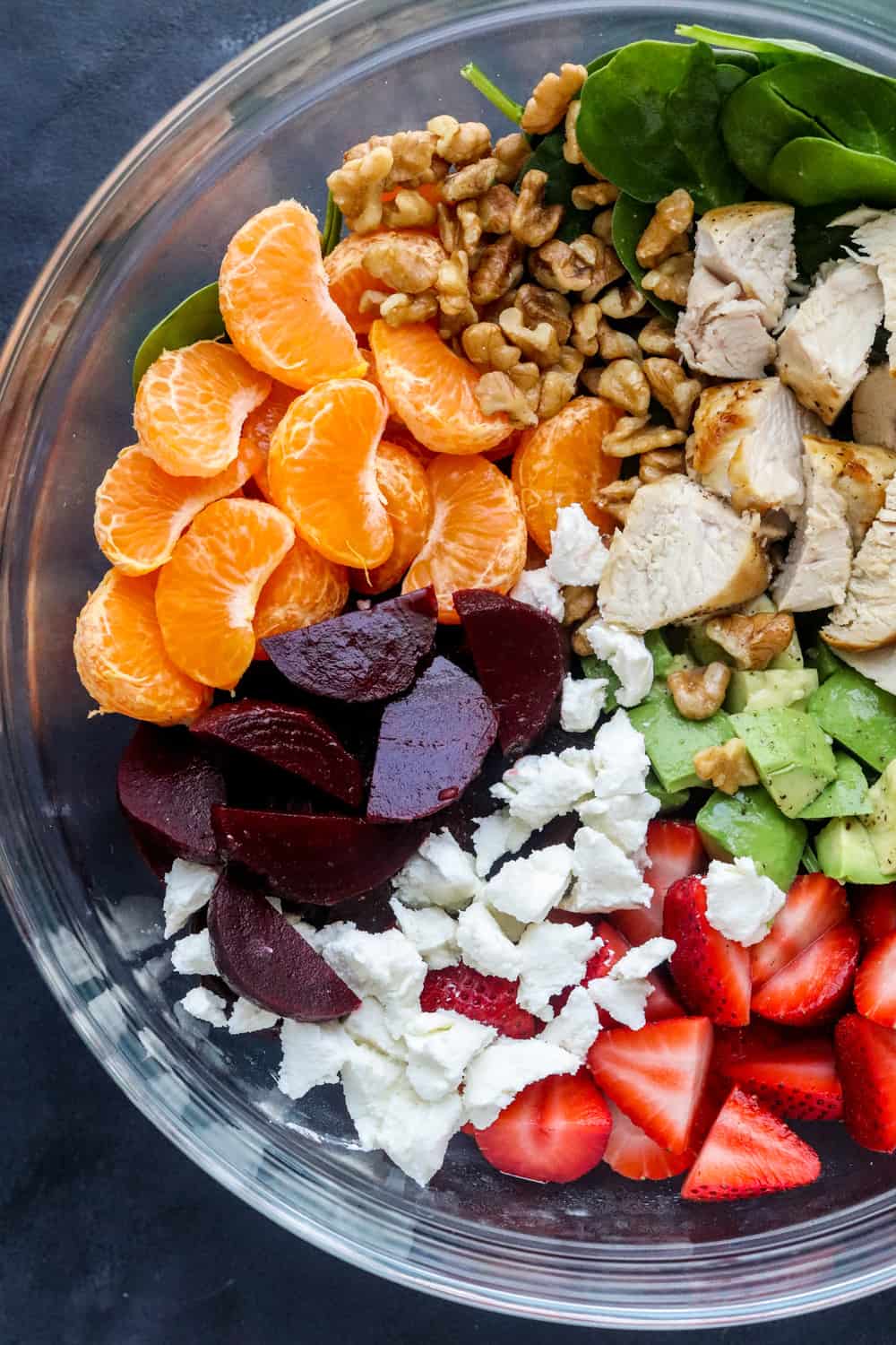 Spinach, clementines, chopped chicken breast, sliced beets, strawberries, avocado and goat cheese in a round glass bowl on a dark blue surface. 