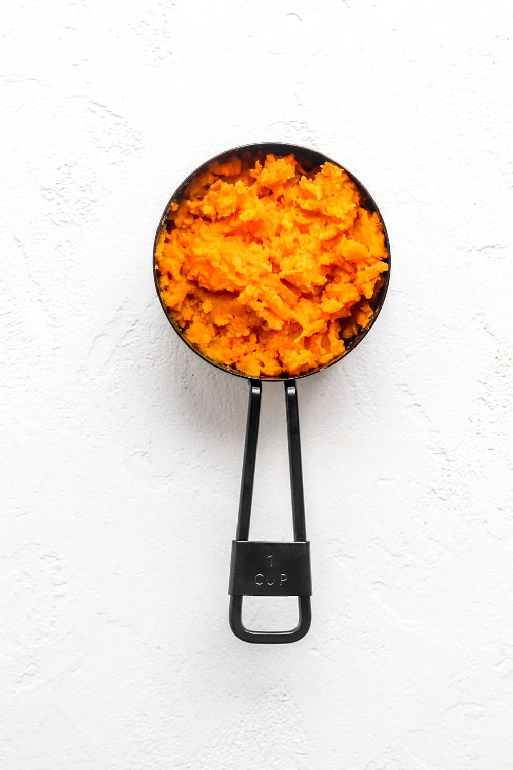 Mashed sweet potato in a black measuring cup on a white surface.