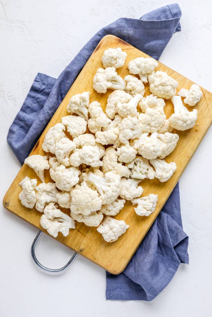 Chopped cauliflower on a wooden cutting board on top of a blue linen. 