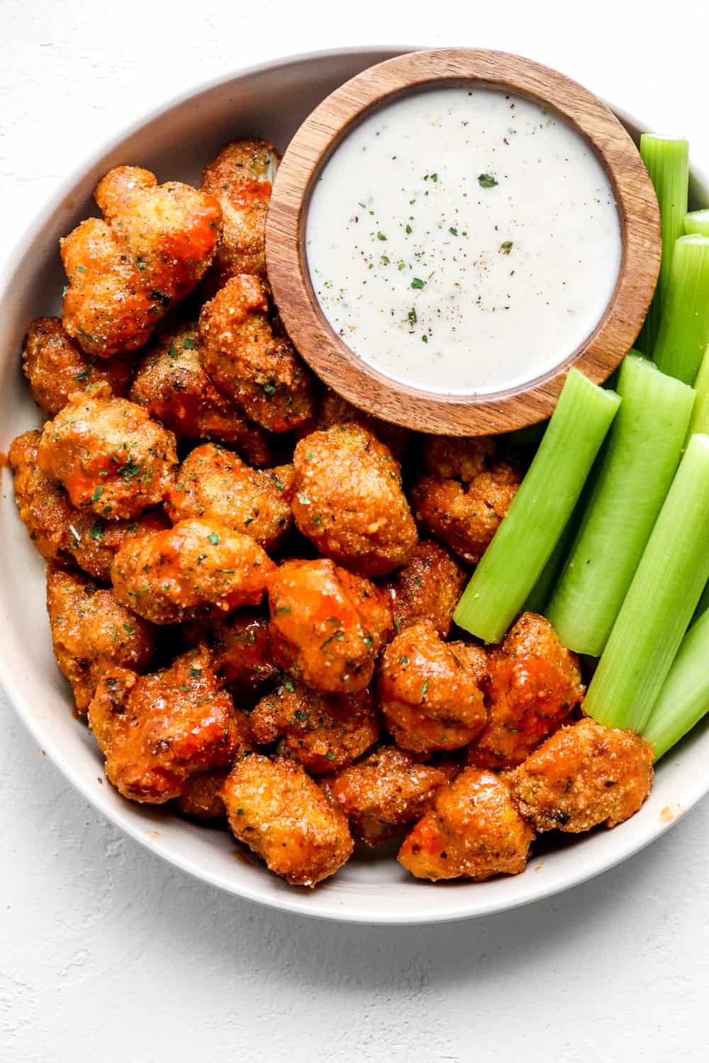 Serving bowl full of buffalo cauliflower wings next to a creamy white sauce with some cut colliery next to it. 