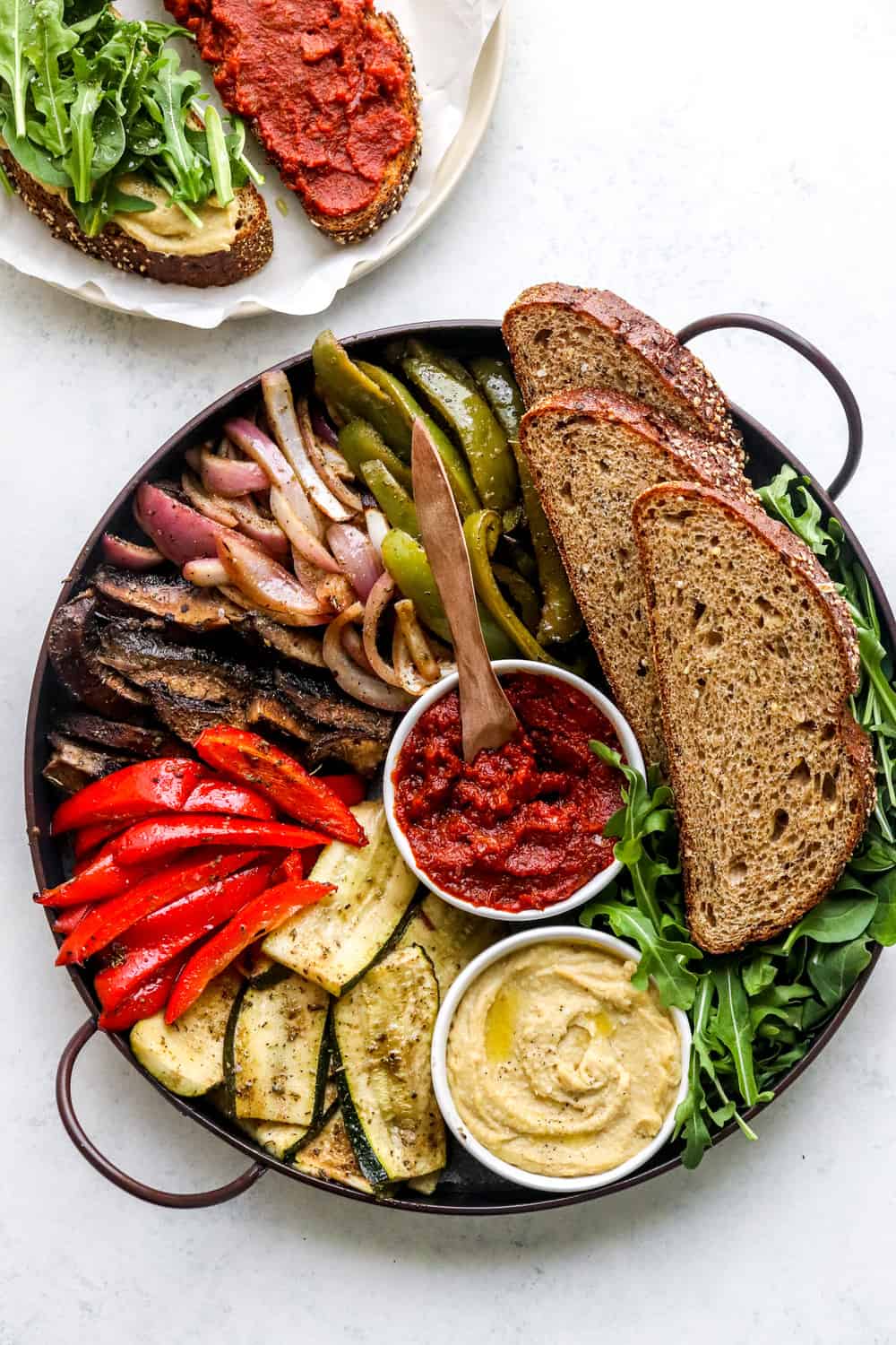 Dark round platter filled with roasted veggies and items for a hummus sandwich with a red tomato spread and hummus in bowls on the platter. 