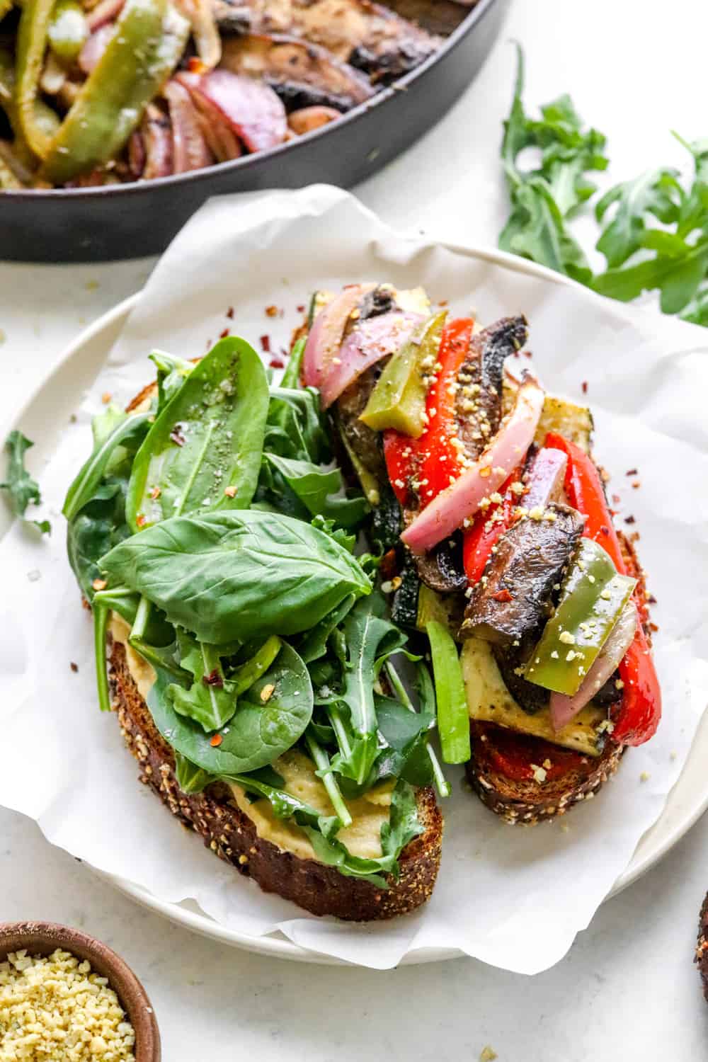 Open faced sandwich filled with greens and toasted bell peppers and mushrooms with vegan parmesan in a brown bowl next to it. 