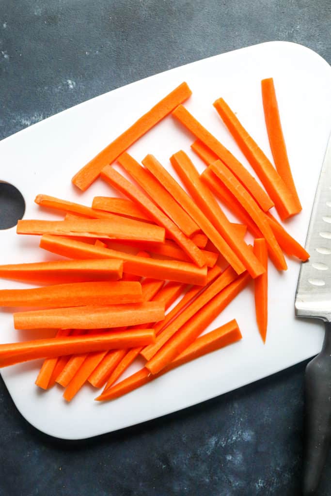 Pile of carrot sticks on a white cutting board with a silver metal knife on the edge of the cutting board.