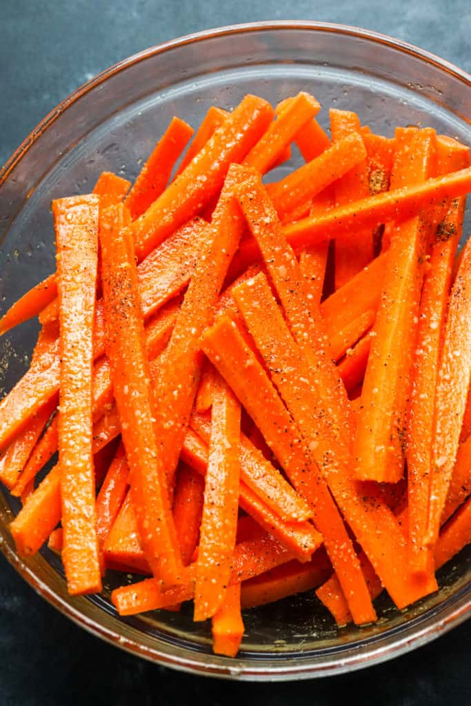 Large round glass mixing bowl filled with seasoned raw carrot fries 
