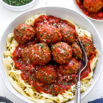 Bowl of linguine topped with meatballs covered in red sauce topped with parsley with a fork cutting into one of the meatballs