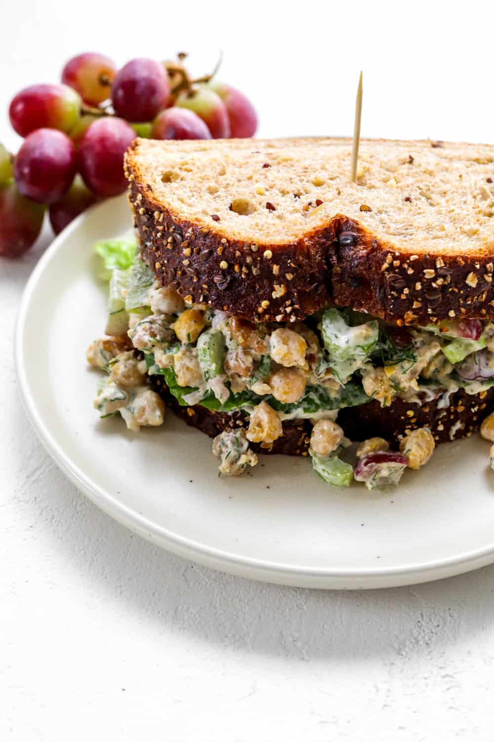 whole grain chickpeas veggie sandwich on a round white plate with a pile of purple grapes behind it.