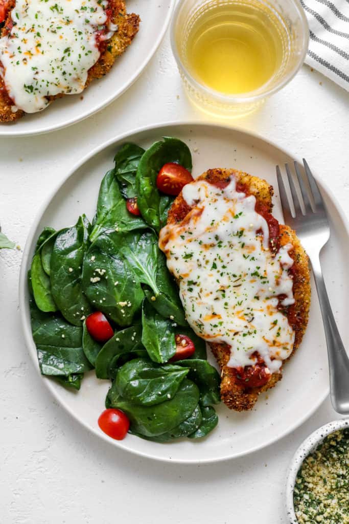 plate of chicken parmesan next to a spinach and tomato salad with a glass cup of white wine behind it