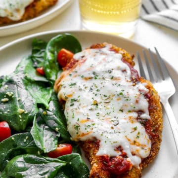 Crispy breaded chicken parmesan on a plate with spinach salad with a form next to the plate and a glass of white wine behind it