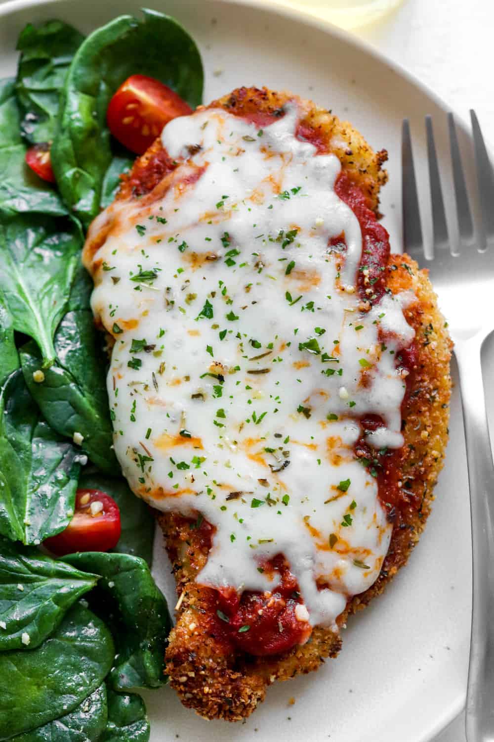 Healthy breaded chicken parmesan on a white plate with spinach and tomato salad next to it and a silver fork on the the plate