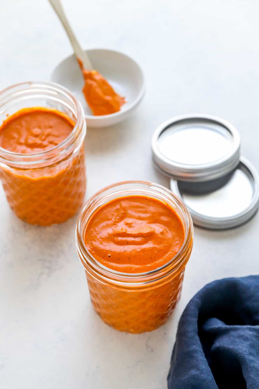 Red pepper romesco sauce in two mason jars with a blue towel next to one and silver lids behind the jars