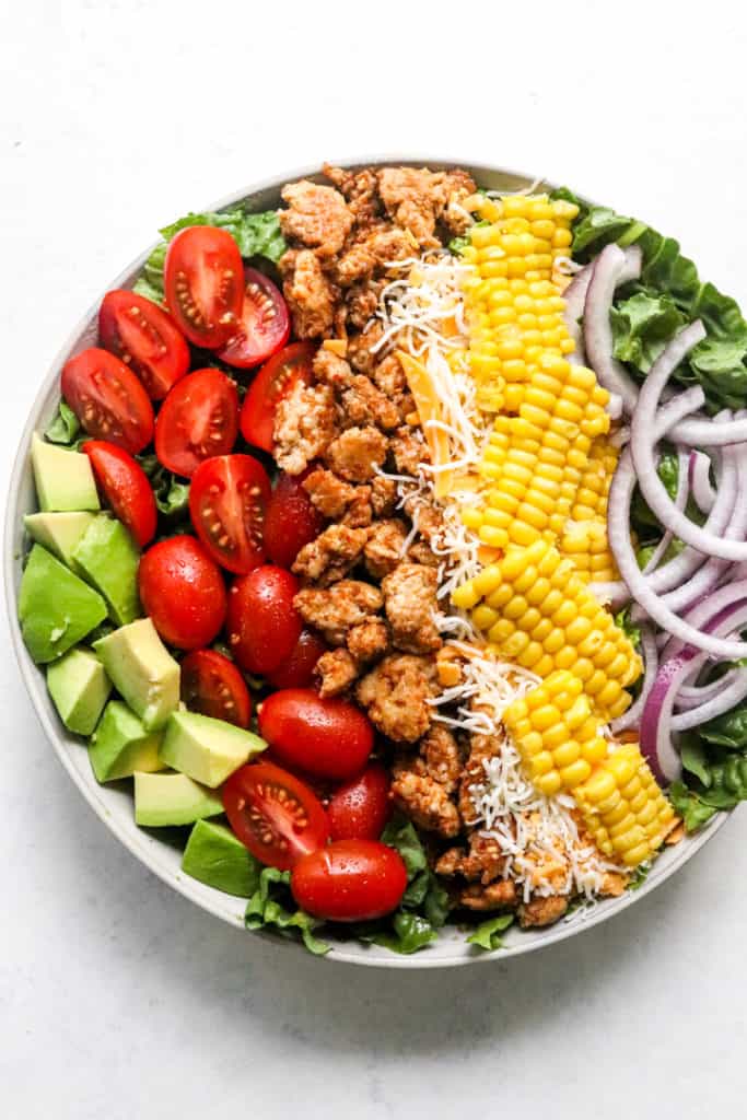 Diced avocado, sliced red tomatoes, cooked chicken, shredded cheese, corn, sliced red onion all piled on top of green lettuce in a round bowl