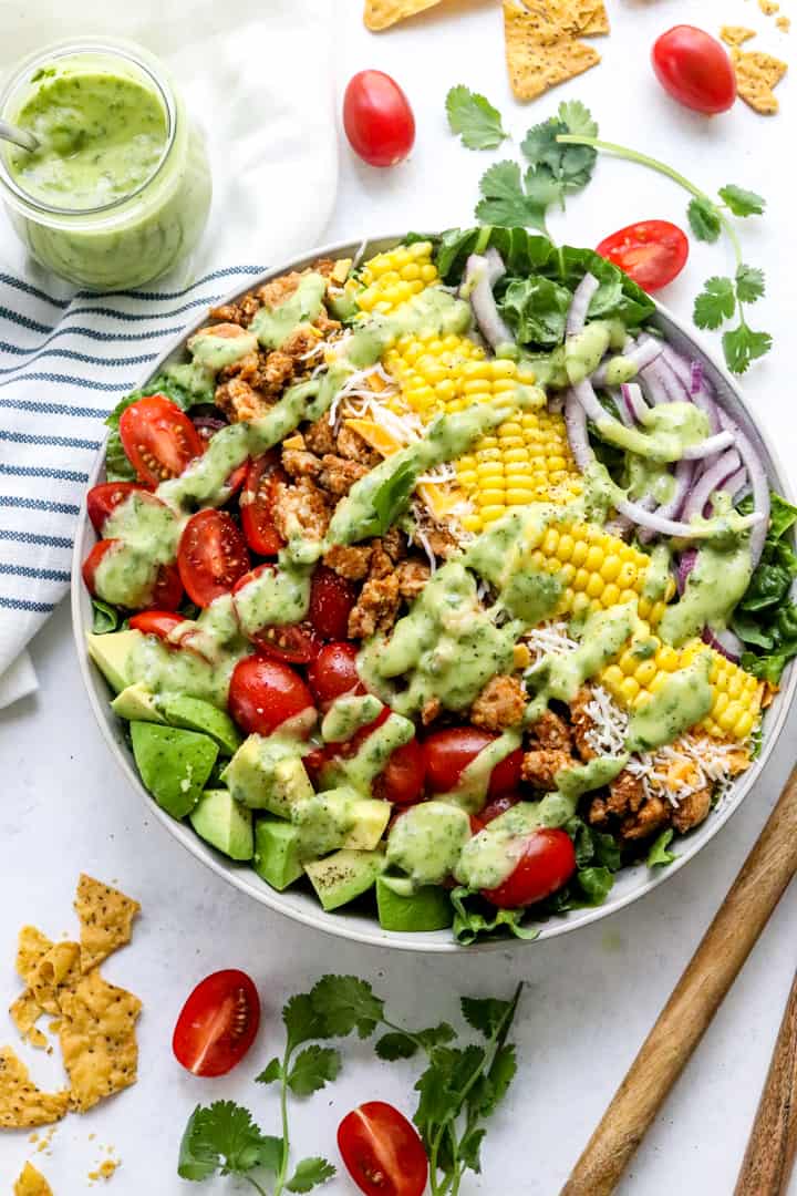 Round bowl filled with lettuce topped with cherry tomato, cooked chicken, corn, red onion with a green dressing on top with a jar of the green dressing behind it. 