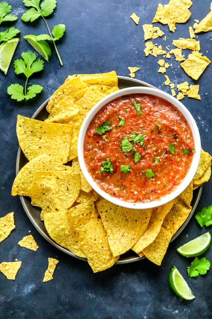 Bowl of tomato salsa on a plate with tortilla chips on it with more crushed behind it and cilantro behind it with lime wedges