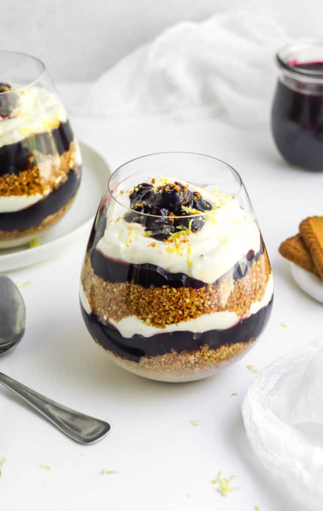 Rounded short glass with layers of crumbled crust, blueberry filling and whipped topping in it with another glass behind it