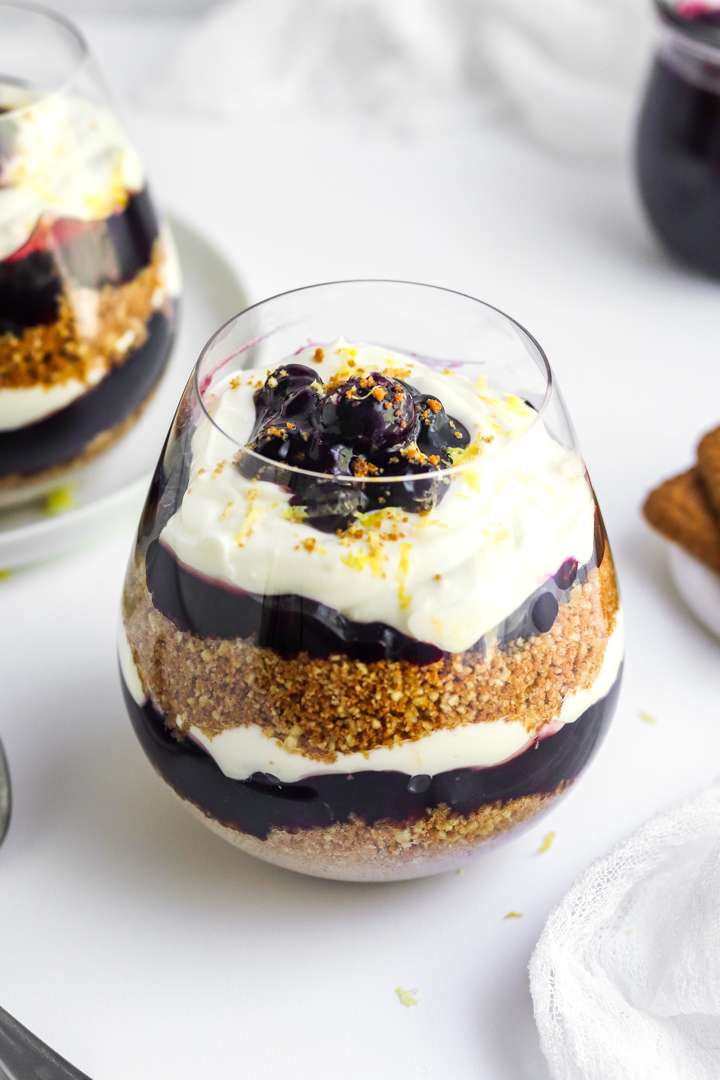 Round glass filled with layered crust, blueberry filling and cheesecake whipped topping with another one behind it. 