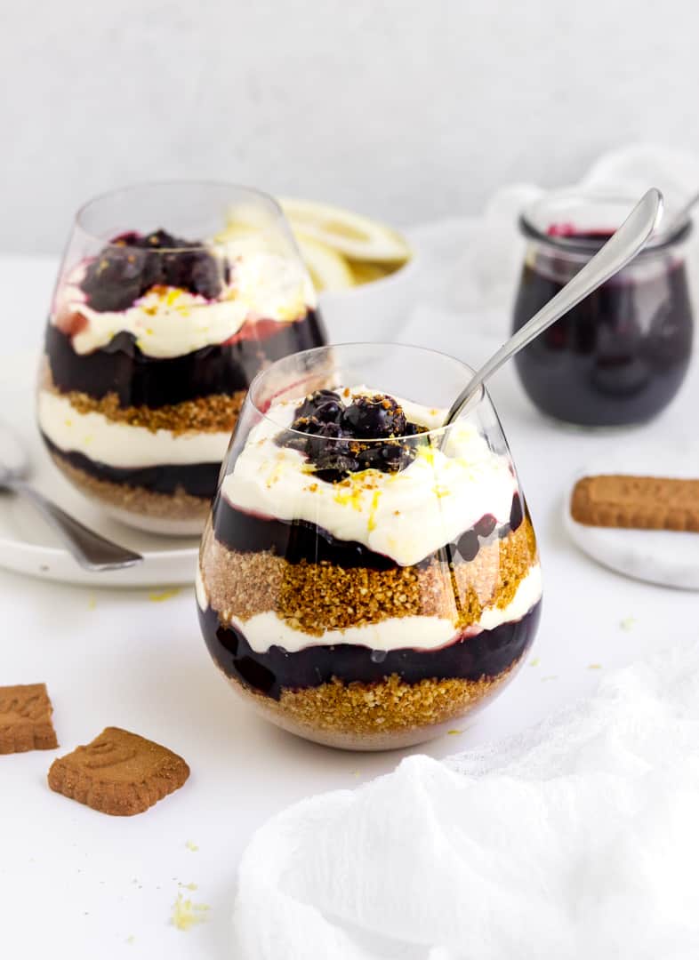 Two rounded glasses with layered cheesecake in them topped with blueberry filling with a spoon in one of the glasses and a broken cookie next to it. 
