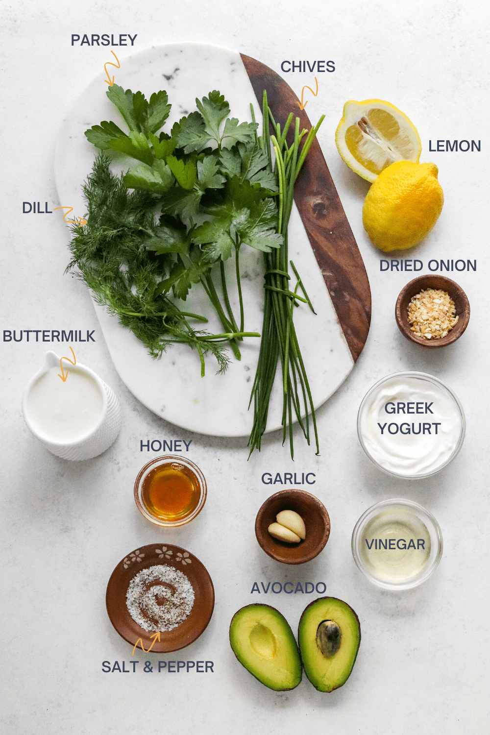 Board with fresh herbs on it with a cut lemon next to it with bowls of buttermilk, yogurt, honey and garlic and vinegar as well as a sliced avocado in front of it. 