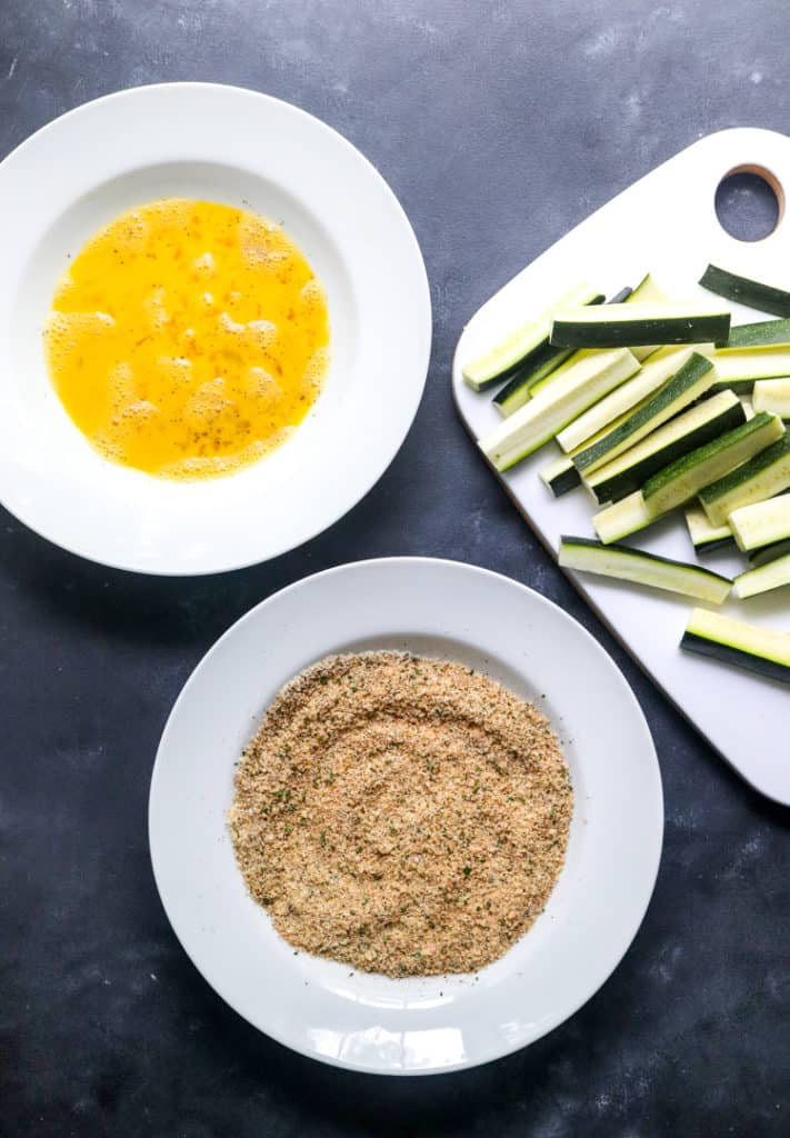 a white bowl filled with whisked egg with another white bowl filled with a  breading mixture and a cutting board with cut zucchini on it next to it