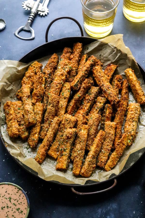 Crispy breaded eggplant fries on a round dark platter with a glass of white wine behind them and a bowl of orange sauce in front of them