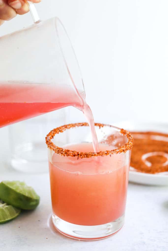  A glass pitcher pouring pink seltzer into a glass filled with grapefruit margarita